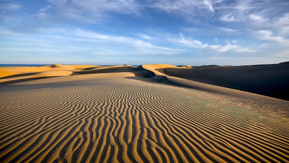 Weitläufige Sanddünen mit markanten Windrillen unter blauem Himmel mit leichten Wolken.